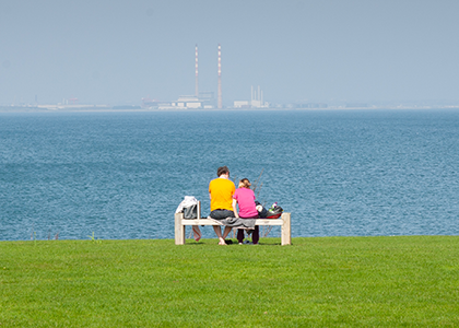 Two young people sit on a bench overlooking the sea. Smokestacks are visible in the background.